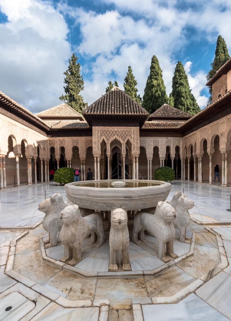Fuente central con los doce leones de mármol en el Patio de los Leones de la Alhambra, rodeada por columnas y arcos de estilo nazarí bajo un cielo parcialmente nublado.